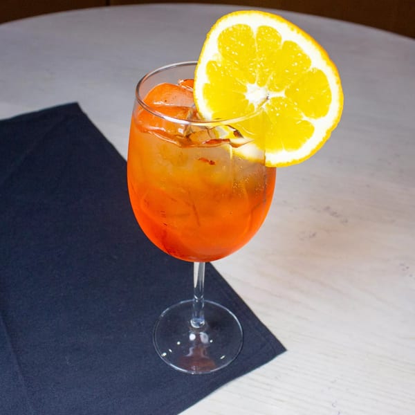 A vibrant orange cocktail with ice and an orange slice garnish, on a white table with a dark napkin.