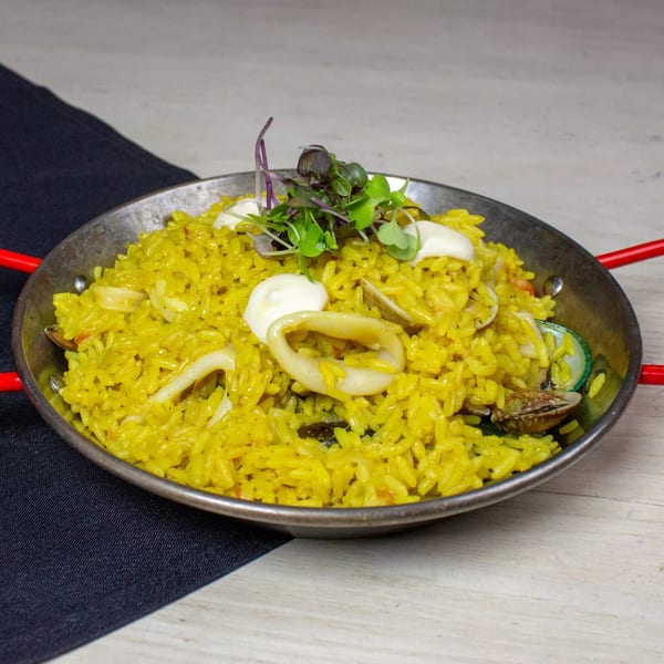 Seafood paella in a metal pan with red handles, garnished with microgreens and white sauce.