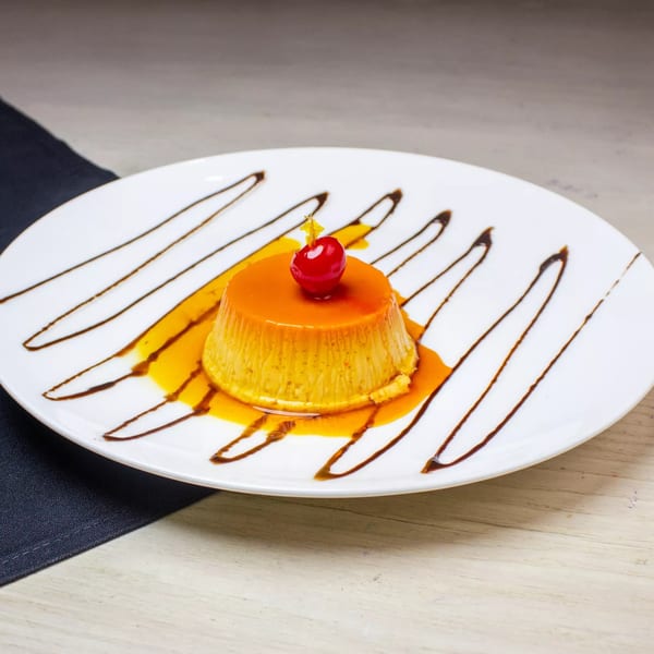 Flan dessert with caramel sauce and a cherry on a white plate with chocolate drizzle.