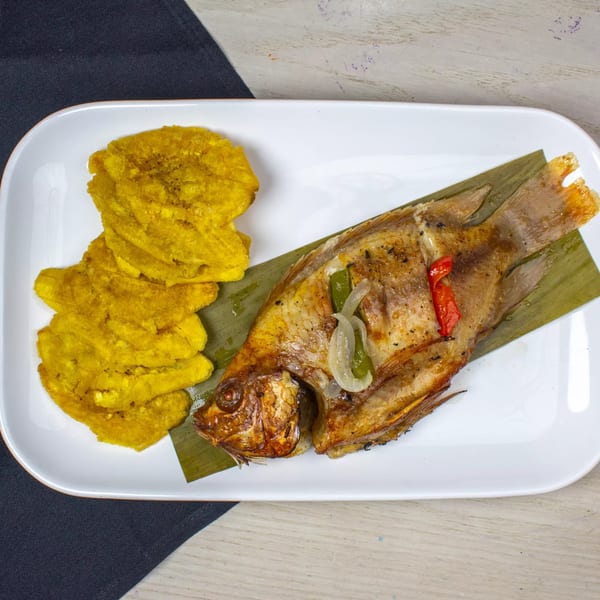 Whole fried fish with peppers and onions on a banana leaf, served with fried plantains.