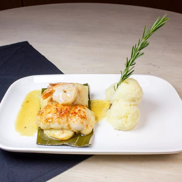 Seafood dish with shrimp and scallops on a banana leaf, mashed potatoes, and rosemary garnish.
